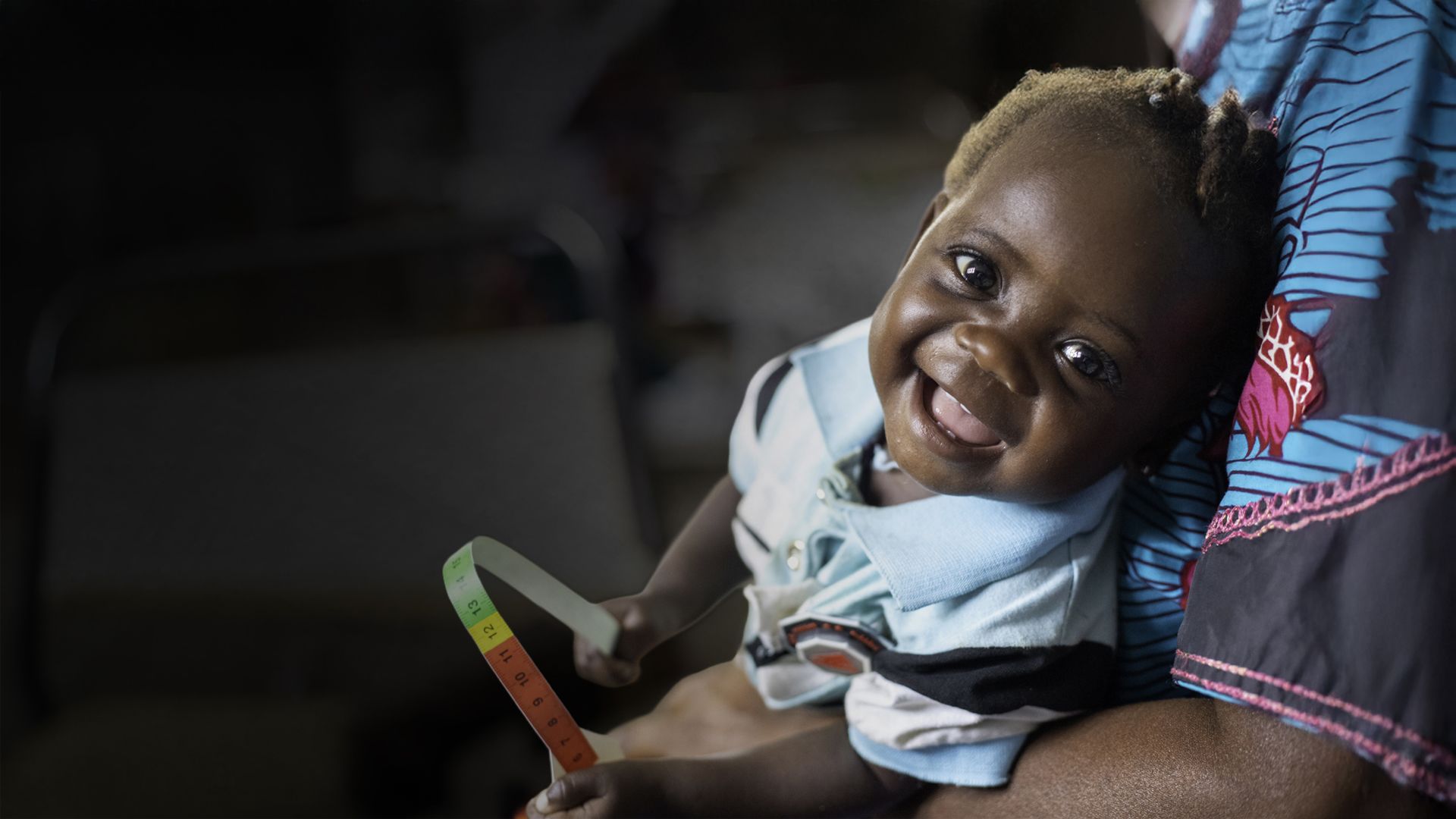 In her mother, Bénita's* arms, Marlène*, 7 months, holds a MUAC band (Brachial perimeter) in her hands